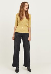Gul, ribbet cardigan med V-udskæring og korte ærmer, parret med mørke, brede jeans og sorte spidse ankelstøvler.
