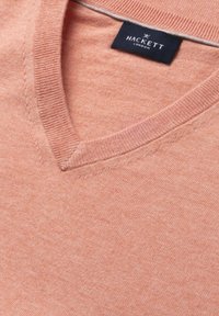 Sweter w kolorze jasnej koralowej z dekoltem w serek, z naszywką Hackett London w kolorze granatowym wszytą wewnątrz kołnierzyka.