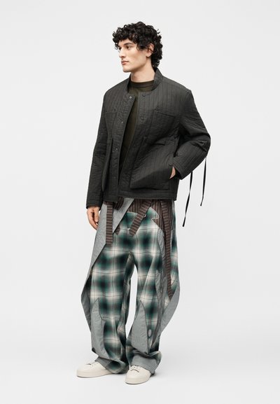 Νέος άνδρας που φοράει μαύρο μπουφάν με quilting, σκούρο πουκάμισο, φαρδιά πράσινη καρό παντελόνια με γκρι υφασμάτινες λεπτομέρειες και λευκά αθλητικά παπούτσια, όρθιος.