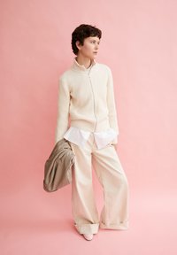 Cremfarbener, gerippter Pullover mit Reißverschluss, über einem weißen Hemd getragen. Weite beige Hosen und eine braun gestreifte Jacke haltend. Sanfter rosa Hintergrund.
