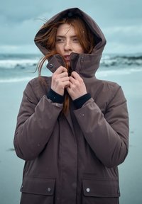 Bruine waterdichte parka met een hoge kraag en capuchon, voorzien van drukknoopsluitingen, zijzakken en een gestructureerde stof, tegen een kustdecor.