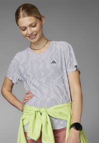 Joven sonriente, con las manos en la cadera, vistiendo una camiseta gris con estampado, una chaqueta verde clara atada a la cintura y un reloj inteligente negro en la muñeca.