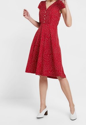 Mujer con un vestido rojo hasta la rodilla con lunares blancos, mangas cortas y zapatos blancos de tacón, de pie frente a un fondo liso.