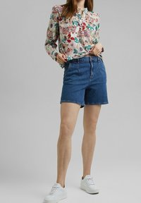 Blouse à manches longues florale avec des couleurs vives, associée à un short en denim bleu. Le mannequin porte des baskets blanches. Tissu doux et coupe décontractée.