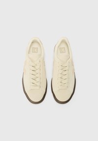 Light beige sneakers with flat laces, reinforced toe cap, and brown rubber sole. Features a textured fabric upper and logo on the tongue.