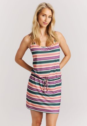 Blonde Frau mit offenem Haar, trägt ein ärmelloses, mehrfarbig gestreiftes Kleid mit Kordelzug in der Taille, posiert mit den Händen hinter dem Rücken.