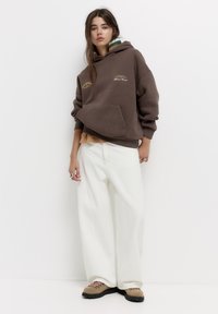 Hnědá oversized mikina s přední kapsou a žlutým textem, doplněná volnými bílými manšestrovými kalhotami a béžovými turistickými botami se šněrováním.