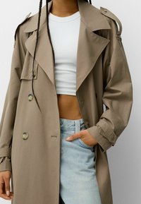 Beige trenchcoat med en bred krage, stora knappar och axelremmar, parat med en vit crop top och ljusblå jeans.
