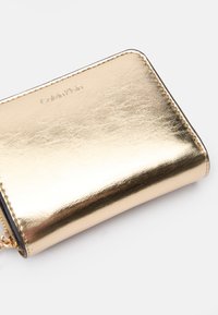 Portefeuille métallique doré avec une texture lisse, fermeture éclair et logo de la marque en relief. Forme rectangulaire avec bords cousus.