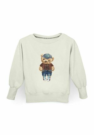 Jasno beżowy sweter z nadrukiem na przodzie przedstawiającym misia w niebieskiej czapce, brązowym swetrze, niebieskich dżinsach i trampkach.