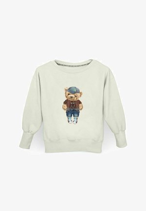 Jasno beżowy sweter z nadrukiem na przodzie przedstawiającym misia w niebieskiej czapce, brązowym swetrze, niebieskich dżinsach i trampkach.