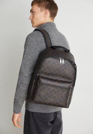 Jeune homme portant un pull gris et un pantalon foncé avec un sac à dos marron à carreaux dans son dos sur fond blanc.
