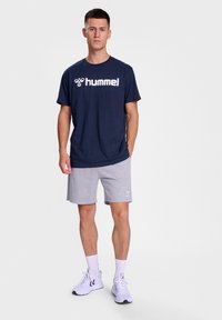 Jūrinės spalvos medvilninis marškinėliai su baltu "hummel" logotipu; šviesiai pilkos šortai; baltos sportinės bateliai; modelis stovi prieš paprastą šviesų foną.