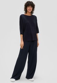 Marineblaue, weit geschnittene Hose kombiniert mit einem lockeren, kurzärmeligen Oberteil. Der Stoff hat eine gerippte Textur. Weiße Slipper runden den Look ab.