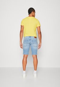 Camiseta amarilla de manga corta con cuello redondo, combinada con shorts de mezclilla azul claro, que presentan un acabado desvaído y mangas enrolladas. Zapatillas blancas visibles.