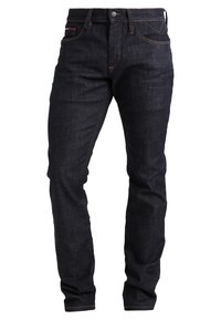 Jean slim bleu foncé avec cinq poches, fermeture par bouton et détail de coutures orange.