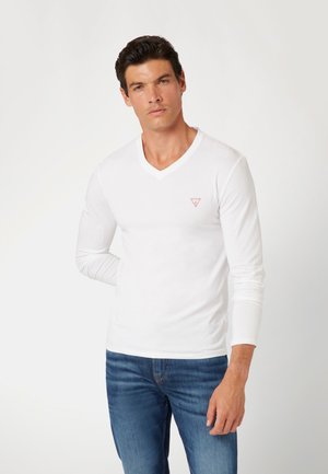 Langærmet hvid v-hals shirt med et lille rødt trekantslogo på venstre bryst, parret med blå jeans. Blød, glat stoftekstur.