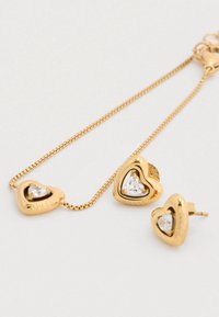 Σετ κοσμημάτων σε χρυσό τόνο με σκουλαρίκια σε σχήμα καρδιάς και ταιριαστό κολιέ, κάθε ένα διακοσμημένο με καθαρό πολύτιμο λίθο και χαραγμένο με τη λέξη "GUESS."