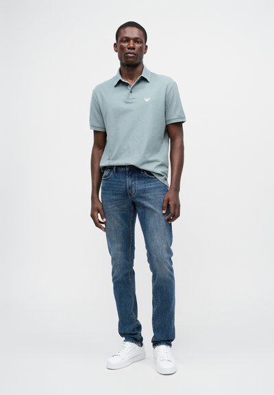 Ανοιχτό μπλε πόλο μπλουζάκι με γιακά, συνδυασμένο με σκούρο μπλε τζιν και λευκά αθλητικά παπούτσια. Το πόλο διαθέτει ένα μικρό λογότυπο στο στήθος.