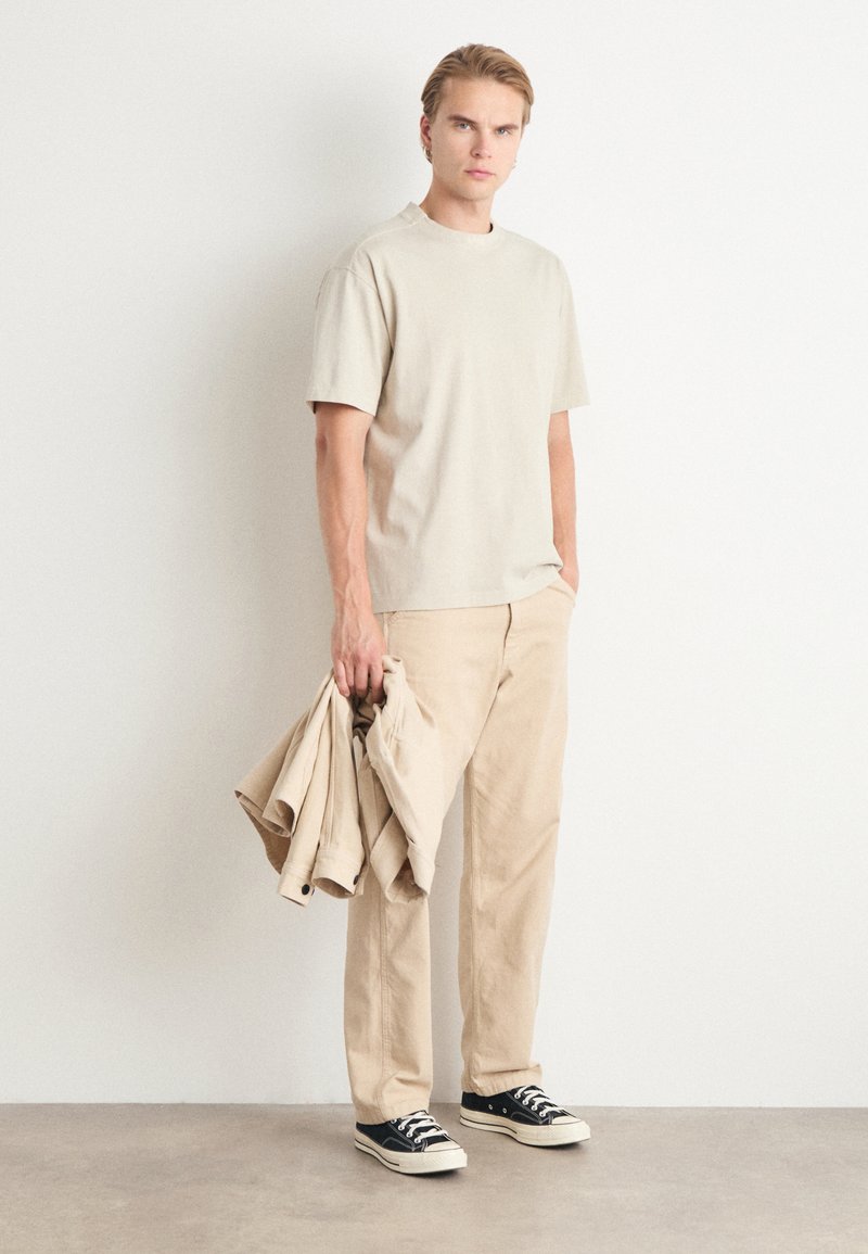 Beige T-shirt met korte mouwen, bijpassende beige broek en zwarte sneakers. Houdt een beige jack vast. Glad materiaal, ontspannen pasvorm, casual ontwerp.