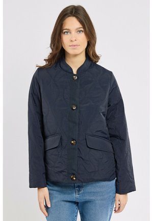Femme portant une veste matelassée bleu marine avec des boutons-pression et des poches à rabat à l'avant, associée à un jean bleu clair, debout devant un fond uni.