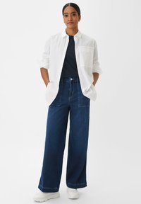 Weißes Hemd mit Knöpfen, dunkelblaues T-Shirt, hochgeschnittene, weit geschnittene dunkelblaue Jeans und weiße Sneakers. Das Outfit hat eine lockere Passform.