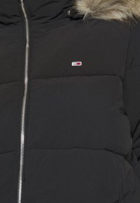 Černá péřová bunda se stříbrným předním zipem, malým červeným, bílým a modrým obdélníkovým logem na levém prsu a světle hnědým kožušinovým lemem na kapuci.