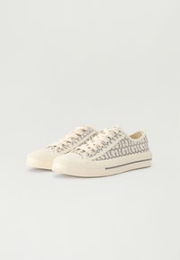 Beige vászon sportcipők, ismétlődő logo mintával, kerek orral, lapos talppal és bézs fűzőkkel, valamint egy kontrasztos, világos színű csíkkal.
