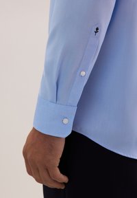 Camicia azzurro chiaro con maniche lunghe, caratterizzata da un polsino con bottoni e un ricamo di dettagli floreali neri vicino alla manica. Tessuto di consistenza liscia.