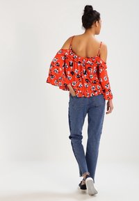 Femme portant un chemisier rouge à fleurs à épaules dénudées, un jean bleu et des baskets noires, debout de dos face à un fond uni.