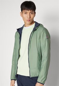 Lys grønn zip-up jakke med mørkeblå hette, elastiske mansjetter og et lite logo-patch på venstre arm. Glatt, lett stoff.