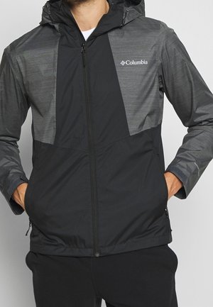 Man wearing a black and gray Columbia hooded jacket with hands in pockets and a front zipper closed.