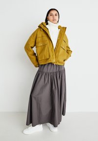 Mosterdgele cropped jas met grote voorzakken, off-white gebreide coltrui en een grijze enkellange rok, gecombineerd met witte sneakers.