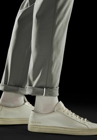 Adidași din piele albă cu numere aurii discrete, purtați cu pantaloni gri deschis cu mansete și șosete albe, pe un fundal întunecat.