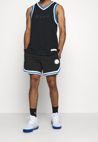 Man som bär en svart Puma ärmlös baskettröja med blå och vit kant, matchande shorts med cirkulärt mönster, svarta strumpor och vita sneakers.