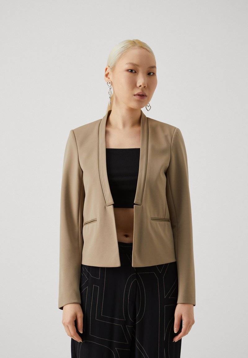Beige cropped blazer in smooth fabric, featuring a shawl collar and two front pockets, paired with a black crop top and wide-leg pants.