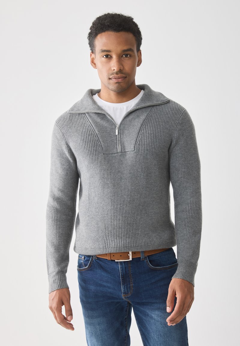 Hall hallitise ja soonilise kudumisega pullover, millel on pooltõmbuv krae, koos siniste teksade ja pruun vööga. Lihtne, tekstuuriga disain ilma mustriteta.