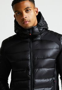 Čierna vypchatá bunda s priliehajúcou kapucňou, so zipsom a textúrovanými panelmi. Má lesklý povrch a jemný červený logo detail na zips.