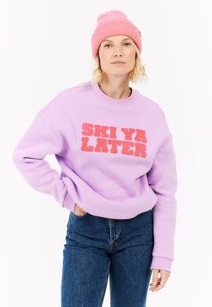 Paarse sweatshirt met "SKI YA LATER" in vet roze letters, geribbelde ronde hals en manchetten. Draagt de sweatshirt met blauwe jeans en een roze gebreide beanie.