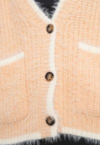 Κοντή κοντή ζακέτα από απαλό νημάτινο ύφασμα σε ροδακινί απόχρωση με λευκή επένδυση. Διαθέτει τέσσερα στρογγυλεμένα καφέ κουμπιά και δύο εμπρόσθιες τσέπες.