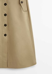Jupe beige longueur genoux avec boutons noirs à l’avant et une seule poche à rabat sur le côté droit sur fond blanc.