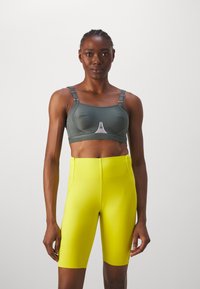 Sostén deportivo gris con tirantes ajustables y un emblema en la parte frontal, combinado con pantalones cortos de ciclismo de talle alto y de color amarillo brillante. Textura suave y ajustada.