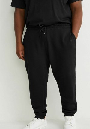 Persoon die zwarte sweatpants met trekkoord en witte sneakers draagt, staand met één hand in de zak tegen een effen achtergrond.