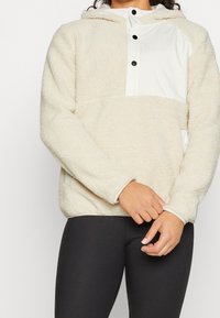 Sudadera de polar beige con un panel blanco en el pecho, botones negros y capucha. Textura suave y corte holgado con mangas largas.