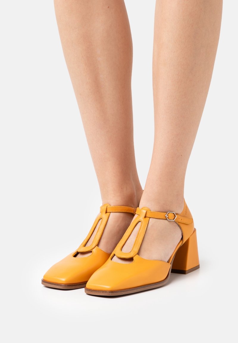 Talons jaunes en cuir avec un design à T, bout carré et un large talon bloc. Texture lisse et détail de sangle de cheville ajustable.