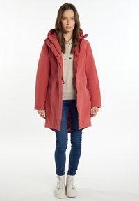Parka rossa di mezza lunghezza con vita regolabile, cappuccio e tasche con zip, indossata sopra una felpa beige e abbinata a jeans blu scuri e sneakers bianche.