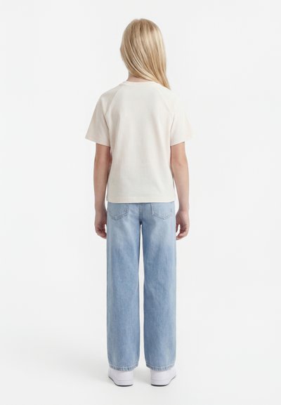 Παιδί με μακριά ξανθά μαλλιά που φοράει λευκό μπλουζάκι με κοντά μανίκια, ανοιχτόχρωμα μπλε φαρδιά τζιν και λευκά αθλητικά παπούτσια, με την πλάτη γυρισμένη σε λευκό φόντο.