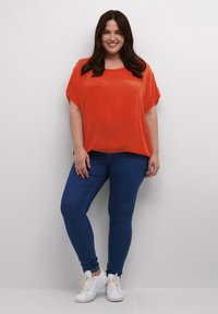 Blouse rouge à manches courtes associée à un jean skinny bleu et des baskets blanches. La blouse a une coupe ample et une texture lisse.