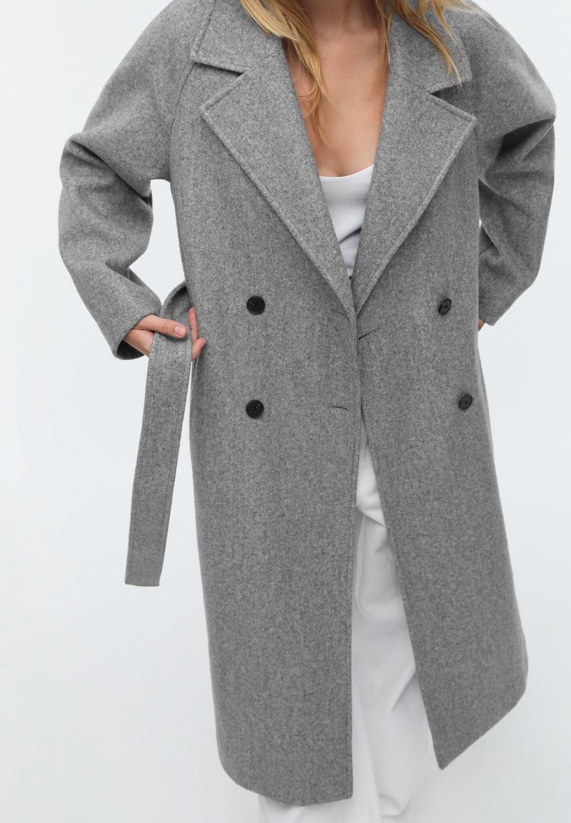 Femme portant un manteau gris en laine à double boutonnage avec des boutons noirs, ajustant la ceinture, sur un pantalon blanc et un haut blanc.