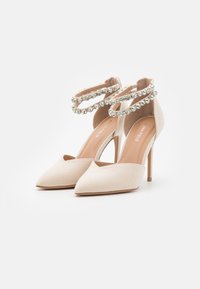 Tacchi alti beige in suede con punta affusolata, adornati da doppi cinturini alla caviglia impreziositi da cristalli. Presentano un design elegante con tacco a stiletto.